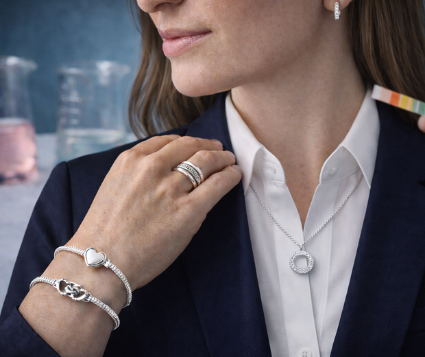 Close-up of a woman in a navy blazer and white buttoned shirt wearing sterling silver earrings, bracelets, ring, and pendant necklace, with blurred lab glassware and a pH test strip in the background.