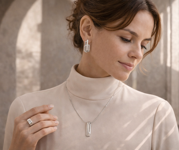 Woman wearing sculptural 925 sterling silver earrings, pendant necklace, and ring in a minimalist architectural setting with soft arch shadows and elegant geometric lines