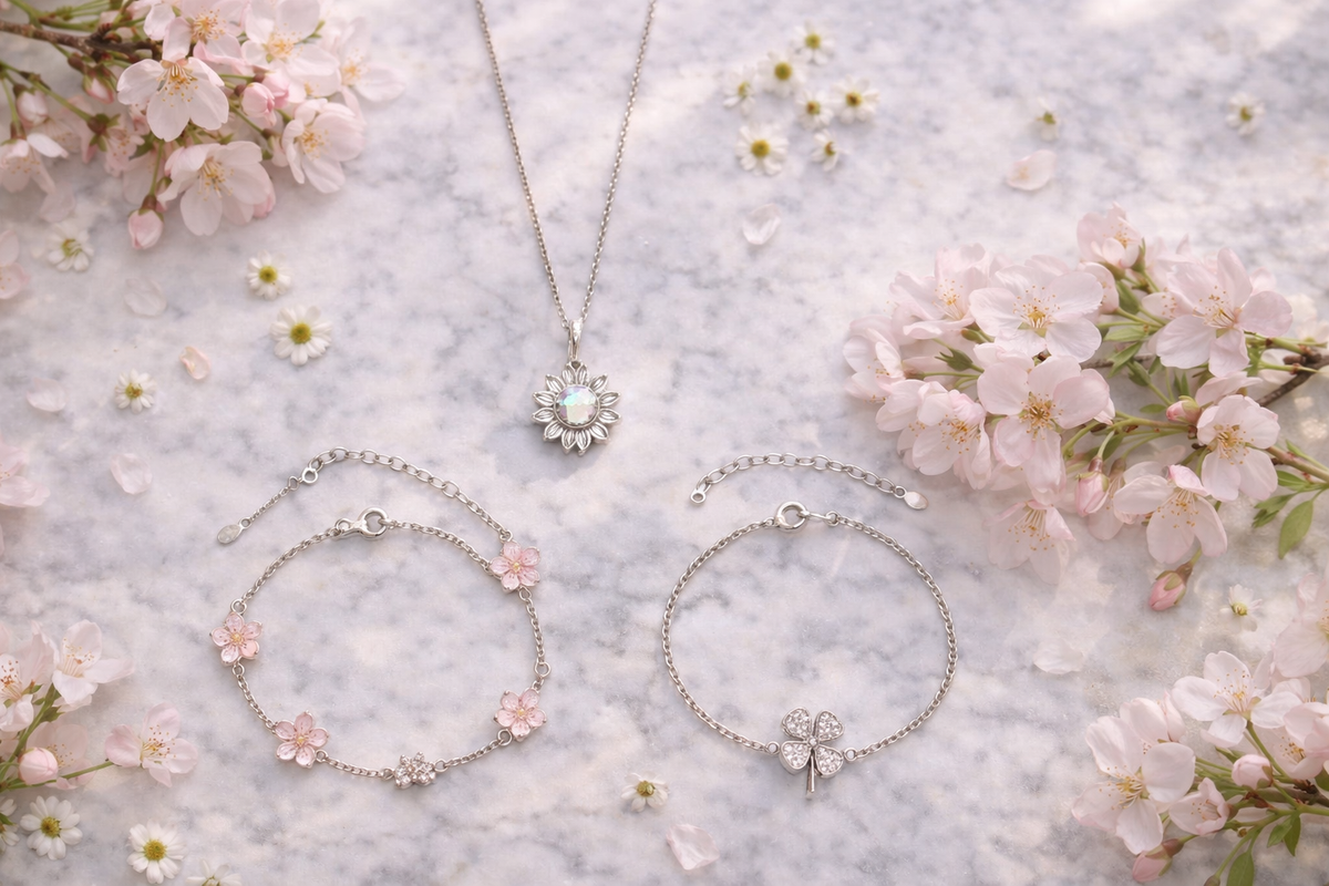 Elegant editorial flat lay of a 925 sterling silver tree of life bracelet, an infinity love bracelet with CZ stones, and a delicate pearl ring, styled with pink roses, soft peonies, green leaves, and a white ribbon gift box in warm natural light.