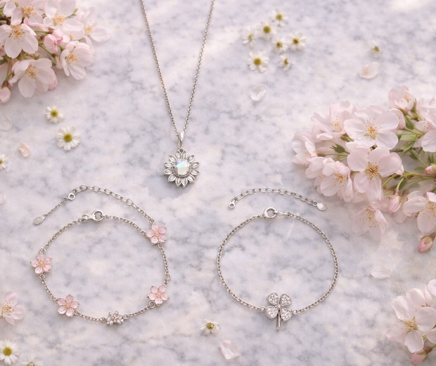 Elegant editorial flat lay of a 925 sterling silver tree of life bracelet, an infinity love bracelet with CZ stones, and a delicate pearl ring, styled with pink roses, soft peonies, green leaves, and a white ribbon gift box in warm natural light.