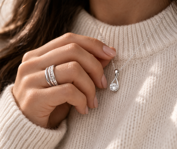 Close-up lifestyle photo of a woman in a cream high-neck sweater gently holding a 925 sterling silver pendant necklace, with stacked silver rings on her finger, warm natural light, and a soft elegant mood.